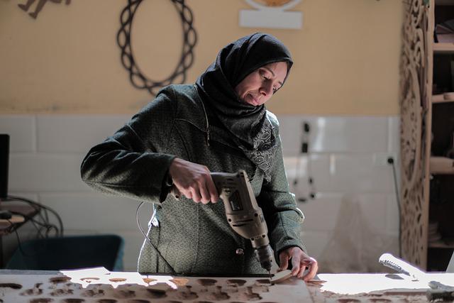woman wearing a hijab stands at a workbench and uses a powered drill to cut holes in disks of wood